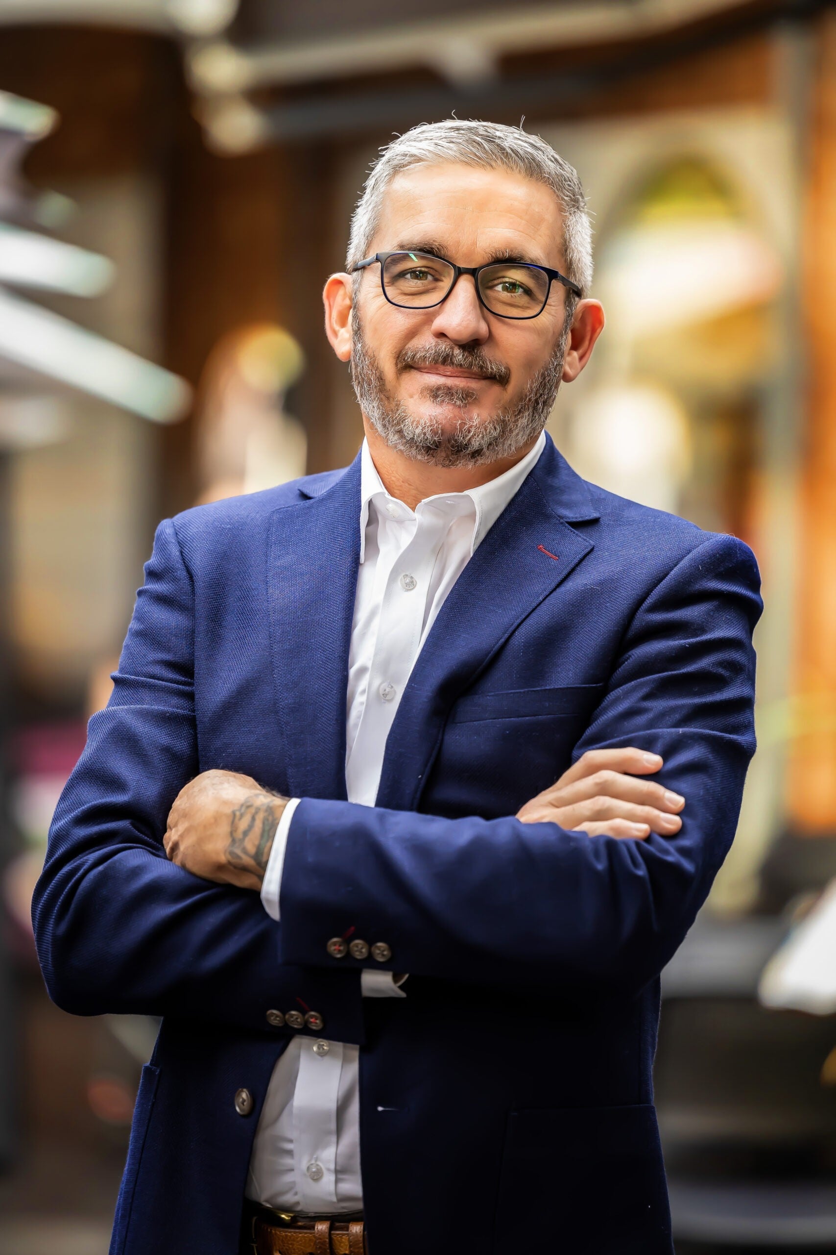 A man with short grey hair, glasses, and a beard smiles confidently with his arms crossed. He is wearing a blue blazer and white shirt, standing in a blurred indoor setting, reminiscent of an independent masseur offering tantric massage Australia.