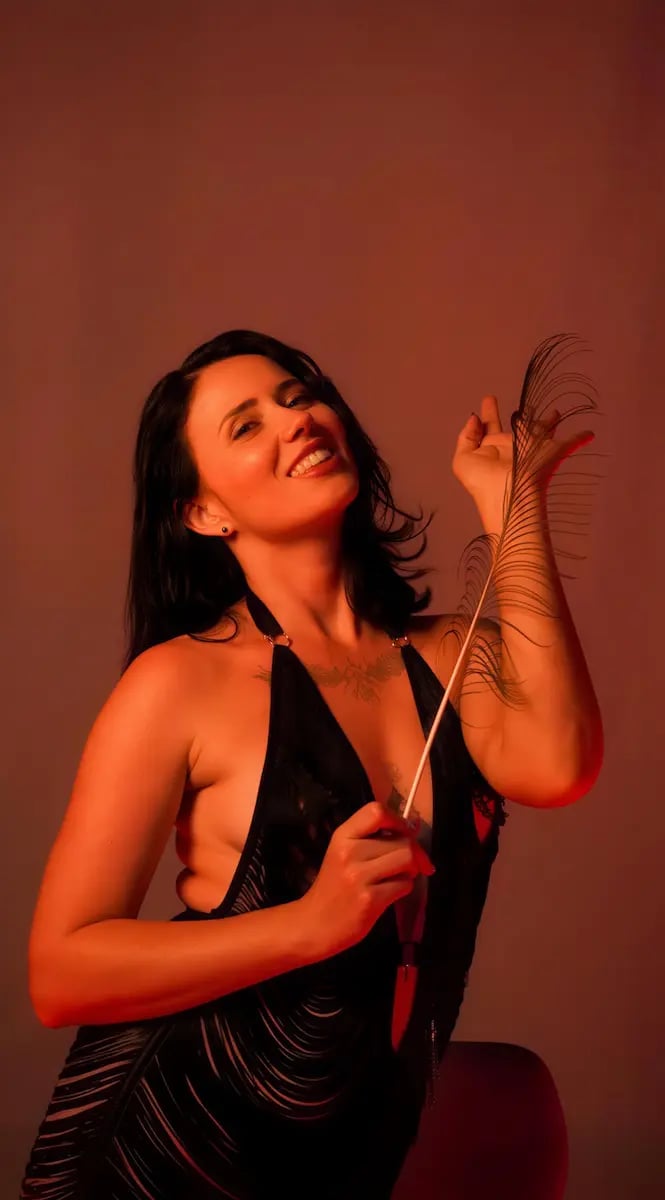 Erotic massage Sunshine Coast, smiling brunette in a black fringed dress holding a feather in warm light Erotic massage Sunshine Coast, smiling brunette in a black fringed dress holding a feather in warm light